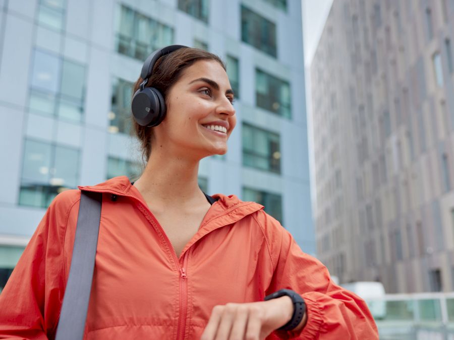Outdoor shot of happy satisfied woman in anorak glad to monitor fitness results on smartwatch listens music from playlist has glad expression poses against blurred background. Sporty lifestyle