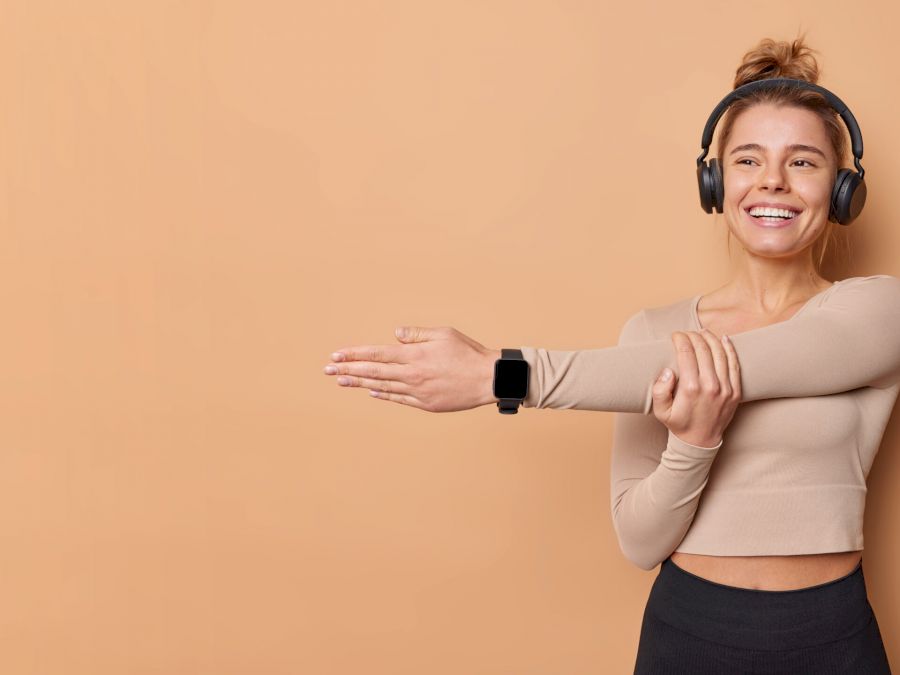 Horizontal shot of active slim healthy woman dressed in activewear stretches arms does training indoor warms up before workout isolated over beige background with blank space for your promotion